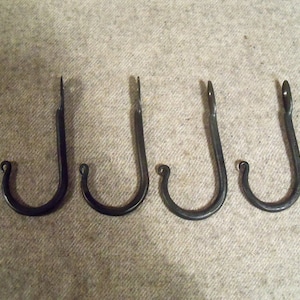 May include: Four hand-forged black metal hooks of varying sizes. Each hook has a curved bottom and a pointed top, ideal for hanging items. The hooks are arranged in a row against a neutral background.
