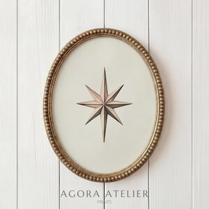May include: An oval framed print featuring a compass rose design. The frame is gold with a beaded edge. The compass rose is in shades of pink and grey on a cream background. The text "AGORA ATELIER PRINTS" is at the bottom.
