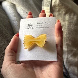 Puede incluir: Un broche hecho a mano con forma de lazo amarillo. El broche está sobre una tarjeta blanca con el texto "Drift Jelly" e información de contacto. El broche se sostiene en una mano con esmalte de uñas rojo.