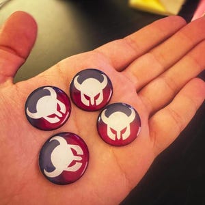 May include: Four round, glossy buttons with a white and red design, held in a hand. The buttons have a dark blue border and a white emblem on a red background. The buttons are approximately 1 inch in diameter.