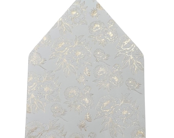 Può includere: Un biglietto triangolare grigio chiaro con un motivo floreale dorato. Il design presenta fiori e foglie dorate dettagliate, creando un aspetto strutturato. La forma del biglietto ricorda una patta di busta.