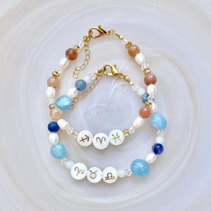 May include: Two beaded bracelets featuring gold-tone lobster clasps and extender chains. The bracelets are strung with white freshwater pearls, clear crystal spacers, and round beads in shades of blue, tan, and white, including zodiac symbol charms.