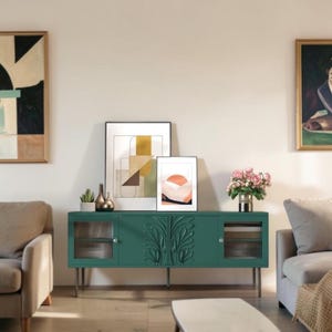May include: A long, dark green sideboard featuring two fluted glass cabinet doors on the sides and a central panel with carved leaf motifs. The unit stands on four thin metal legs and holds two framed abstract prints and a small vase of pink flowers.