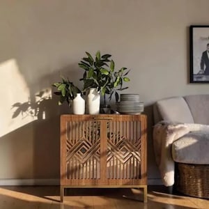 May include: A wooden cabinet with geometric door designs and gold-colored legs. The cabinet is adorned with white vases holding green plants, and a stack of plates. A framed picture and a sofa are in the background.
