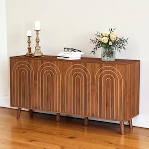 May include: A wooden cabinet with a warm brown finish, featuring a repeating arched design in a lighter tone. The cabinet has four doors and sits on short, tapered legs. Two brass candlesticks, a stack of books, glasses, and a vase of flowers adorn the top.