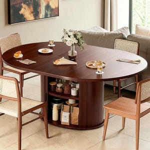 May include: A dark brown oval dining table with a central pedestal base featuring open storage shelves. The table is surrounded by four wooden chairs with woven cane backrests. The tabletop is set with small plates, napkins, and a glass vase of flowers.