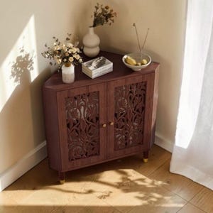 May include: A dark wood corner cabinet featuring two doors with intricate laser-cut floral patterns, small gold-toned knobs, and four short legs with gold-toned feet. The top surface holds a white vase with flowers, a stack of books, and a bowl.