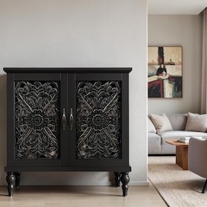 May include: A black cabinet featuring two doors with intricate, carved floral and leaf patterns. The cabinet stands on four dark, rounded, turned legs and includes two small, metallic vertical handles centered between the two doors.
