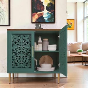 May include: A teal cabinet with a natural wood top and four light-colored tapered legs. The cabinet features two doors with intricate carved floral patterns and a vertical gold handle. Inside, two shelves hold white ceramic bowls and containers.