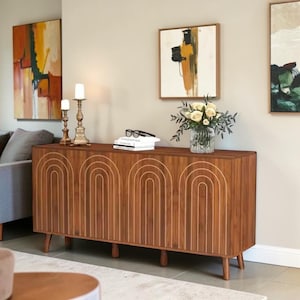 May include: A rectangular wooden sideboard featuring four cabinet doors with carved vertical arch patterns. The piece stands on five tapered legs and has a smooth brown finish. On top sit two brass candle holders, a stack of books, and a glass vase.