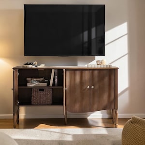 May include: A dark brown wooden TV stand with a ribbed door design and two silver knobs. The stand has two shelves, one with books and a woven basket. A large black TV is mounted above the stand.