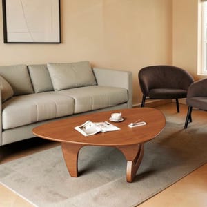 May include: A low-profile, triangular wooden coffee table with rounded edges and curved legs. The tabletop features a smooth, medium-brown wood grain finish. An open magazine and a small white ceramic teacup sit on the surface of the table.