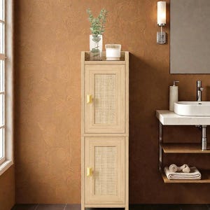 May include: A light wood cabinet with two doors, each featuring a woven cane panel. The cabinet has gold-colored handles and is topped with a glass vase, a candle, and greenery. The cabinet is in a bathroom setting.