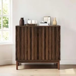 May include: A dark brown wooden cabinet with vertical grooved detailing on the front. The cabinet has a rectangular shape and sits on four short legs. The top of the cabinet displays decorative items, including a vase, mugs, and a tray.