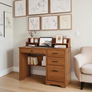 May include: A wooden desk featuring a central drawer, three stacked drawers on the right side, and an open shelf with books. The desk surface holds a laptop, a small vase with dried flowers, a cup, and a small framed picture on a hutch.