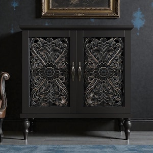 May include: A black cabinet with ornate, carved floral designs on the doors. The cabinet has a rectangular shape, four short legs, and antique-style handles. The background features a framed picture and a dark wall.