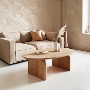 May include: A light-colored, modern living room scene featuring a beige sofa with several throw pillows. A wooden oval coffee table sits in front of the sofa. A vase with dried flowers and a book are on the table. The walls are a light beige color.