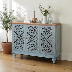 May include: A light blue cabinet with three doors featuring intricate cut-out designs. The cabinet has a wooden top and sits on short, rounded legs. A potted plant and vase of flowers are on top.