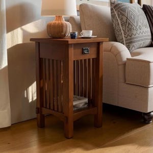 May include: A rectangular wooden end table with a warm brown finish, featuring a single drawer with a dark metal pull handle, vertical slat sides, and an open lower shelf. The table has four sturdy legs and a flat top surface for storage.