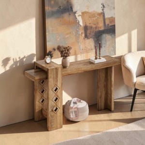 May include: A light brown wooden console table with a rectangular top and a tiered side section. The left support features three diamond-shaped cutouts. The table has a visible wood grain texture and a sturdy, block-like construction design.