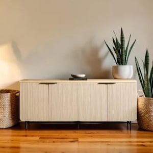 May include: A light-colored wooden sideboard with four doors and black handles. The cabinet has a textured, vertical-lined design. Two potted snake plants and a woven basket are on either side of the cabinet. A small bowl sits on top.
