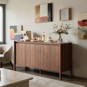 May include: A long, rectangular wooden sideboard with four vertical cabinet doors and slender legs. On top sits a beige espresso machine, a small bowl of fruit, a glass bottle, a clear container, and a glass vase filled with white flowers.