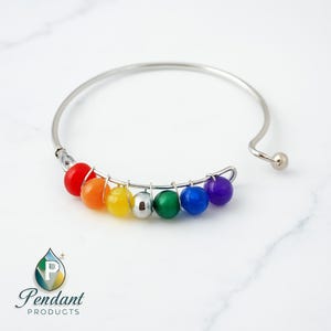 Op de afbeelding: Een zilverkleurige armband met een rij kleurrijke kralen in een regenboogpatroon. De kralen zijn rood, oranje, geel, zilver, groen, blauw en paars. De armband heeft een eenvoudig, open ontwerp met een kleine bal aan het uiteinde.