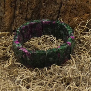 May include: A stretch bracelet made of rectangular green and pink stones. The bracelet is resting on a bed of dried, light brown plant fibers, with a dark brown, textured background.