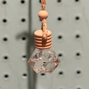 Puede incluir: Un ambientador de coche de cristal transparente, facetado, con una tapa de madera y una cuerda trenzada para colgarlo. El ambientador tiene forma de diamante y está lleno de un líquido transparente. La tapa de madera es de color marrón claro.