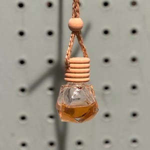May include: A hanging car air freshener with a clear, faceted glass bottle containing a light amber liquid. The bottle has a wooden cap and is suspended by a braided tan cord with a wooden bead.