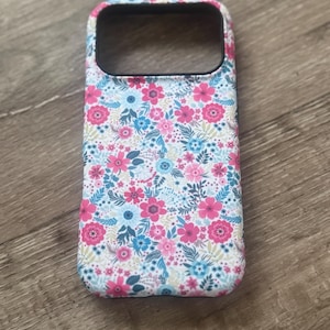 May include: A floral phone case featuring a pattern of pink, blue, and white flowers and green leaves. The case has a black border around the camera cutout and is set against a wooden surface.