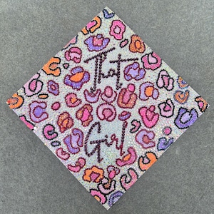 May include: A square graduation cap with a leopard print design in pink, orange, and purple. The words "That Girl" are spelled out in small, sparkling beads. The cap is set against a gray background.