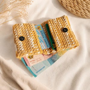 May include: A small, rectangular, crocheted wallet in mustard yellow and white yarn. The wallet features two flaps, each secured with a silver-toned metal snap button. Inside, it holds several Indonesian identification cards and paper banknotes.