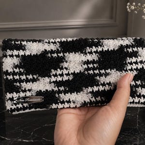 May include: A rectangular crochet clutch featuring a black and white houndstooth pattern. The accessory includes a small silver-toned metal tag on the bottom left corner with the engraved text Ness Handmade in a cursive script font.