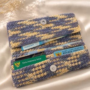 May include: A rectangular crochet wallet featuring a variegated pattern of blue and yellow yarn. The wallet has a fold-over flap secured by a silver magnetic snap button and contains multiple slots holding various identification cards.