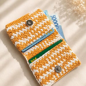 May include: A rectangular crochet wallet featuring a repeating mustard yellow and white geometric pattern. The wallet has a fold-over flap secured by a silver-toned magnetic snap button and contains a blue identification card and paper currency.