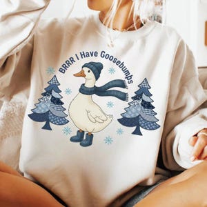 May include: Cream-colored sweatshirt featuring a cartoon goose wearing a blue hat, scarf, and boots. The goose is between two blue and white patterned Christmas trees. Text above the goose reads "BRRR I Have Goosebumps" with snowflakes.