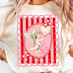 以下が含まれることがあります： クリーム色のTシャツ。ピンクと赤のデザインで、ハート型の風船を持ったケルビムが描かれています。ピンクの楕円形のフレームと赤いストライプで囲まれています。「love」の文字が筆記体で書かれています。