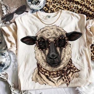 May include: A cream-colored t-shirt featuring a graphic illustration of a sheep wearing brown leopard-print glasses and a matching leopard-print neck scarf. The sheep has a dark face and textured wool, centered on the front of the garment.
