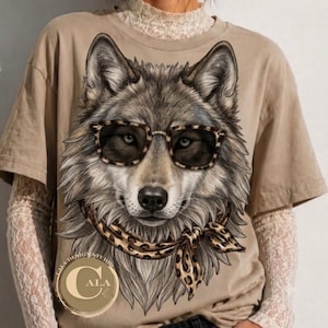 Puede incluir: Una camiseta color canela con una ilustración gráfica de un lobo con gafas de sol de estampado de leopardo y un pañuelo a juego. El lobo tiene pelaje gris y marrón detallado. Se ve un logotipo circular con el texto CALA DESIGN STUDIO.