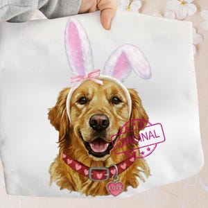 May include: A white tote bag featuring a golden retriever wearing bunny ears and a red collar with heart accents. The dog has a pink heart-shaped tag that says "LOVE". A pink stamp that says "ORIGINAL" is on the bag.