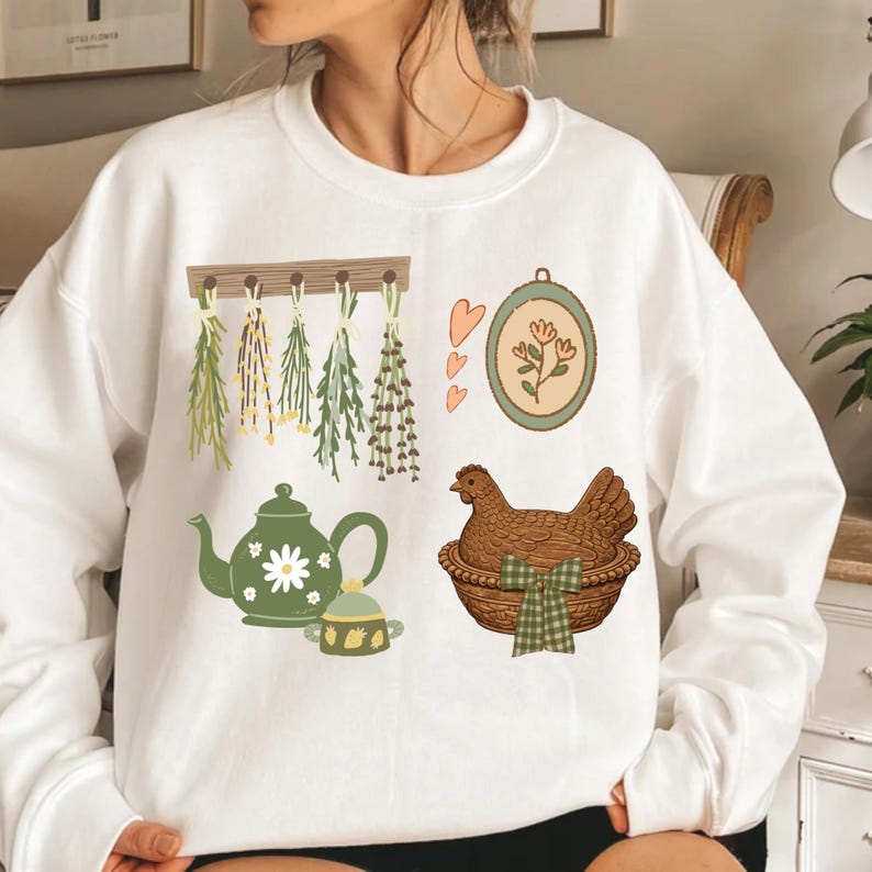 May include: White sweatshirt featuring a farmhouse-themed design. The design includes a green teapot with a daisy, a hen in a basket, hanging herbs, and a framed floral illustration. The overall aesthetic is rustic and charming.