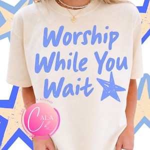 Peut inclure: T-shirt crème avec l'inscription bleue "Worship While You Wait" et une étoile bleue. Le t-shirt a des manches courtes et une coupe décontractée. Un logo est visible dans le coin inférieur gauche.
