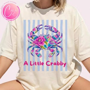 May include: Cream-colored t-shirt featuring a floral crab design in shades of pink, blue, and purple. The text "A Little Crabby" is printed in pink below the crab. The background has light blue vertical stripes.