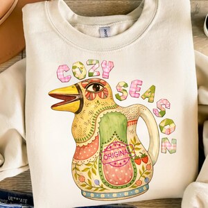 May include: Cream-coloured sweatshirt featuring a bird-shaped pitcher design. The pitcher has a yellow beak, green and pink detailing, and the words "COZY SEASON" in pink. A small "ORIGINAL" stamp is also visible on the design.