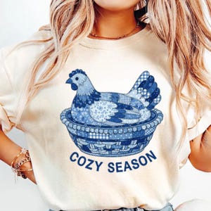 Könnte beinhalten: Cremefarbenes Sweatshirt mit einem blauen Patchwork-Hühnerdesign in einer dekorativen Schüssel. Das Huhn besteht aus verschiedenen blauen Mustern. Die Worte "COZY SEASON" sind blau unter der Schüssel aufgedruckt. Das Sweatshirt hat lange Ärmel.
