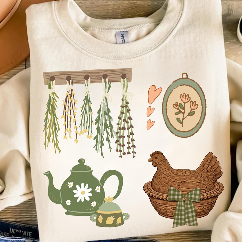 May include: Cream-colored sweatshirt featuring a collection of illustrated elements. The design includes hanging herbs, a green teapot with a daisy, a chicken in a basket, and a floral framed picture. The overall theme is a rustic, farmhouse aesthetic.
