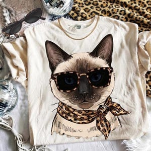 May include: A cream-colored t-shirt featuring a graphic illustration of a Siamese cat wearing leopard print sunglasses and a matching leopard print neck scarf. The cat has blue eyes and dark brown fur on its ears and face.