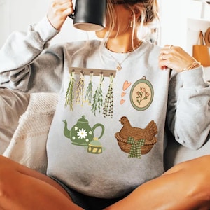 May include: A light gray sweatshirt featuring several nature-themed illustrations. The design includes a green teapot with a daisy, a chicken in a basket, hanging herbs, and a floral embroidery hoop. The overall theme is rustic and cozy.