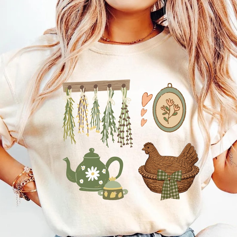 May include: Cream-colored t-shirt featuring a collection of illustrated elements. Includes a teapot, herbs, a chicken in a basket, and a floral framed design. The design has a rustic, farmhouse aesthetic.